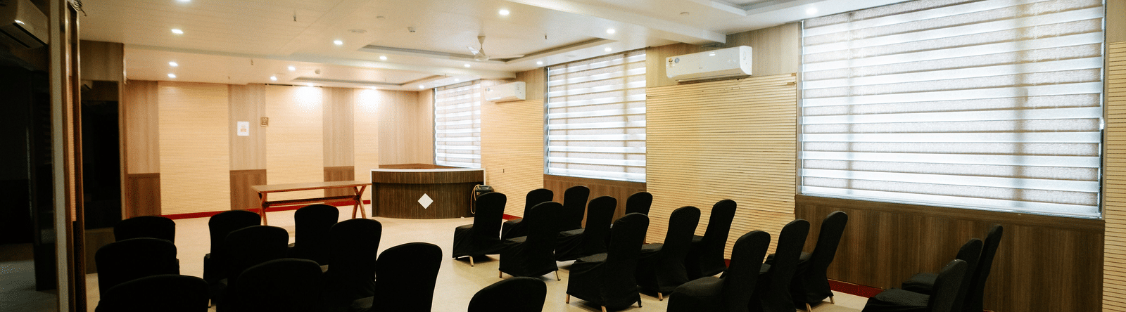 Closer view of the rows of black upholstered chairs in the event space, showing the light wood floors and curtained windows at Daksh The Madhuvan Suites, Dwarka.