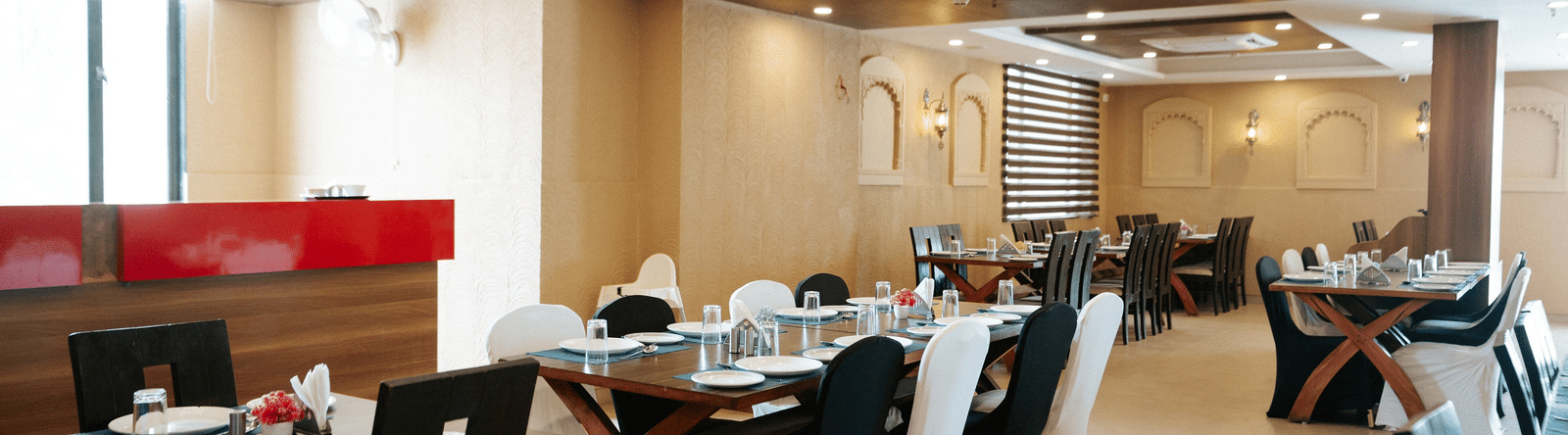 Detail of a restaurant table setting with a blue runner, plates, and dark chairs set against a textured wall with arches at Daksh The Madhuvan Suites, Dwarka.