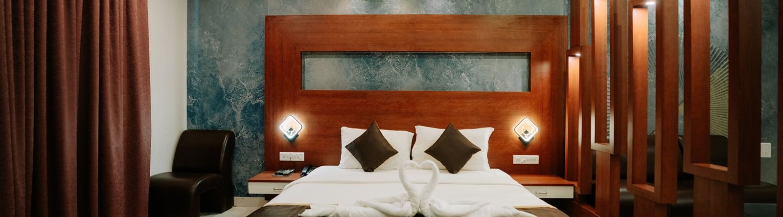 Close-up of the double bed flanked by wooden slats and warm bedside lighting, highlighting the dark wooden headboard at Daksh The Madhuvan Suites, Dwarka.