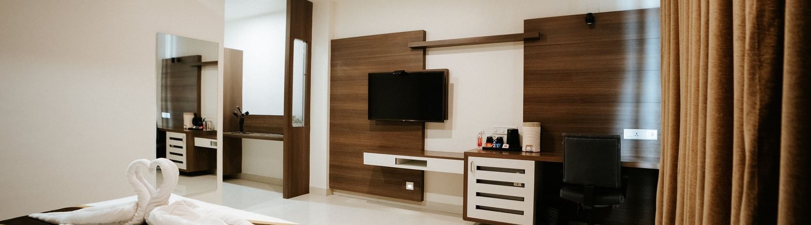 Panoramic view of the upscale hotel room, showing the bed, dark wood furniture, media console, and rich brown curtains at Daksh The Madhuvan Suites, Dwarka.