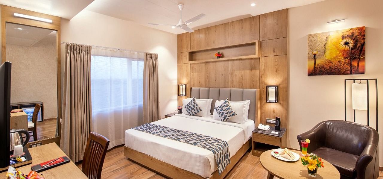 A hotel room showcasing a double bed with white and grey bedding, a wooden bedside table with a lamp, a work desk area with a chair and television, and a small round table with a brown armchair, complemented by wooden flooring at Lords Inn Motikhavdi