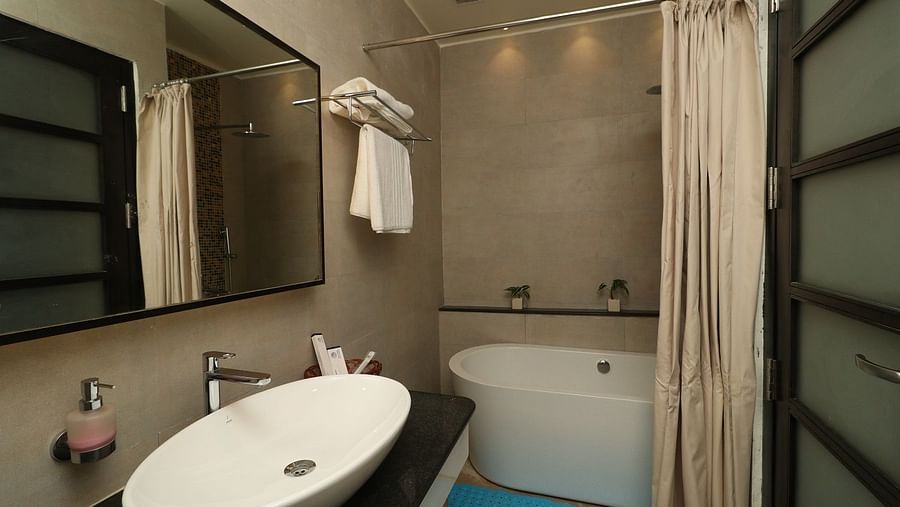 A hotel bathroom with a modern sink and vanity, a mirror, a toilet, and a bathtub with a shower curtain, with a blue bathmat on the floor - Sea Hills Hotels, Sri Vijaya Puram (Port Blair)
