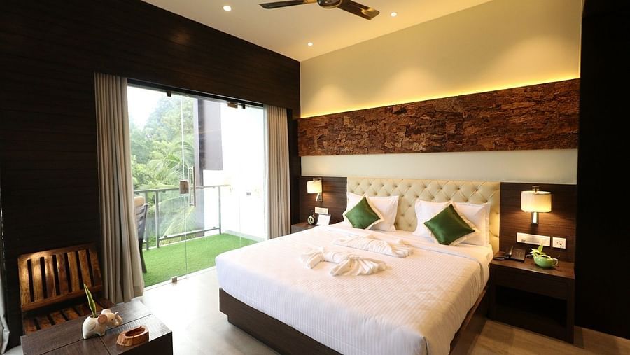 A modern hotel bedroom with a large white bed, bedside tables with lamps, and a view of the outside through a large glass door, featuring a ceiling fan and textured wall - Sea Hills Hotels, Sri Vijaya Puram (Port Blair)