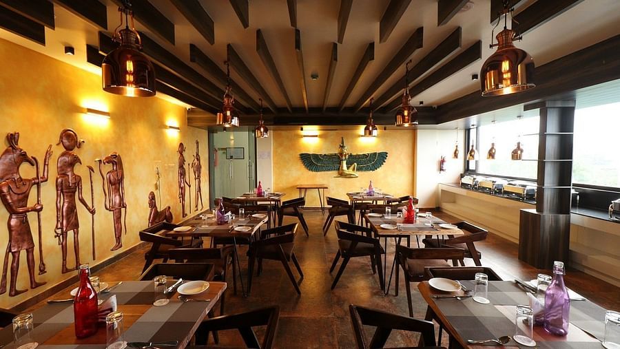 A long shot of the restaurant interior, emphasizing the rows of wooden tables and chairs, the exposed wooden beams on the ceiling, and the artistic wall embellishments - Sea Hills Hotels, Sri Vijaya Puram (Port Blair)