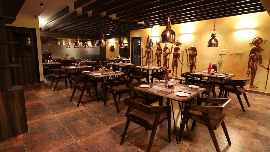 A central perspective of a restaurant dining area, showing multiple set tables with wooden chairs, warm lighting from hanging fixtures, and distinct wall decorations - Sea Hills Hotels, Sri Vijaya Puram (Port Blair)