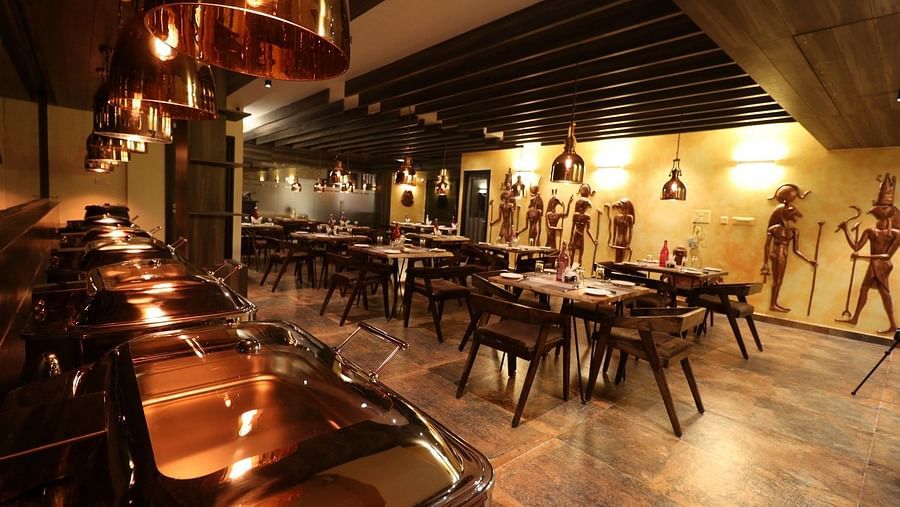 A warm and inviting restaurant interior with a bar or service counter on the left, many wooden tables and chairs, and decorative elements on the walls - Sea Hills Hotels, Sri Vijaya Puram (Port Blair)