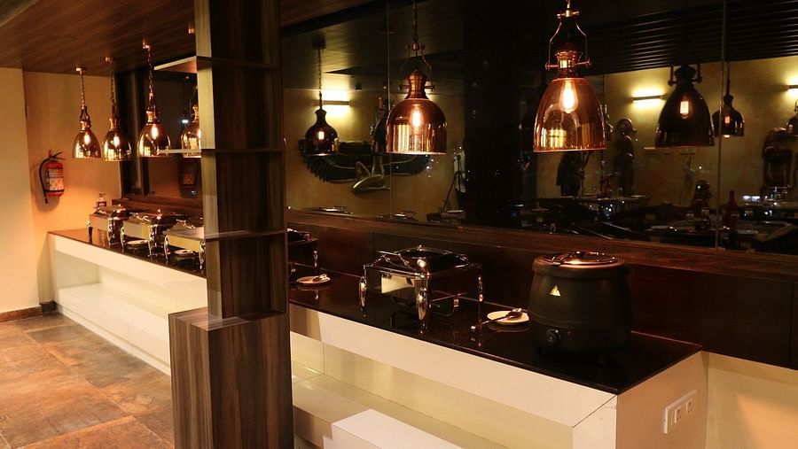 A close-up view of a section of the restaurant, focusing on a wooden counter or service area with decorative lighting and a glimpse of the dining space beyond - Sea Hills Hotels, Sri Vijaya Puram (Port Blair)