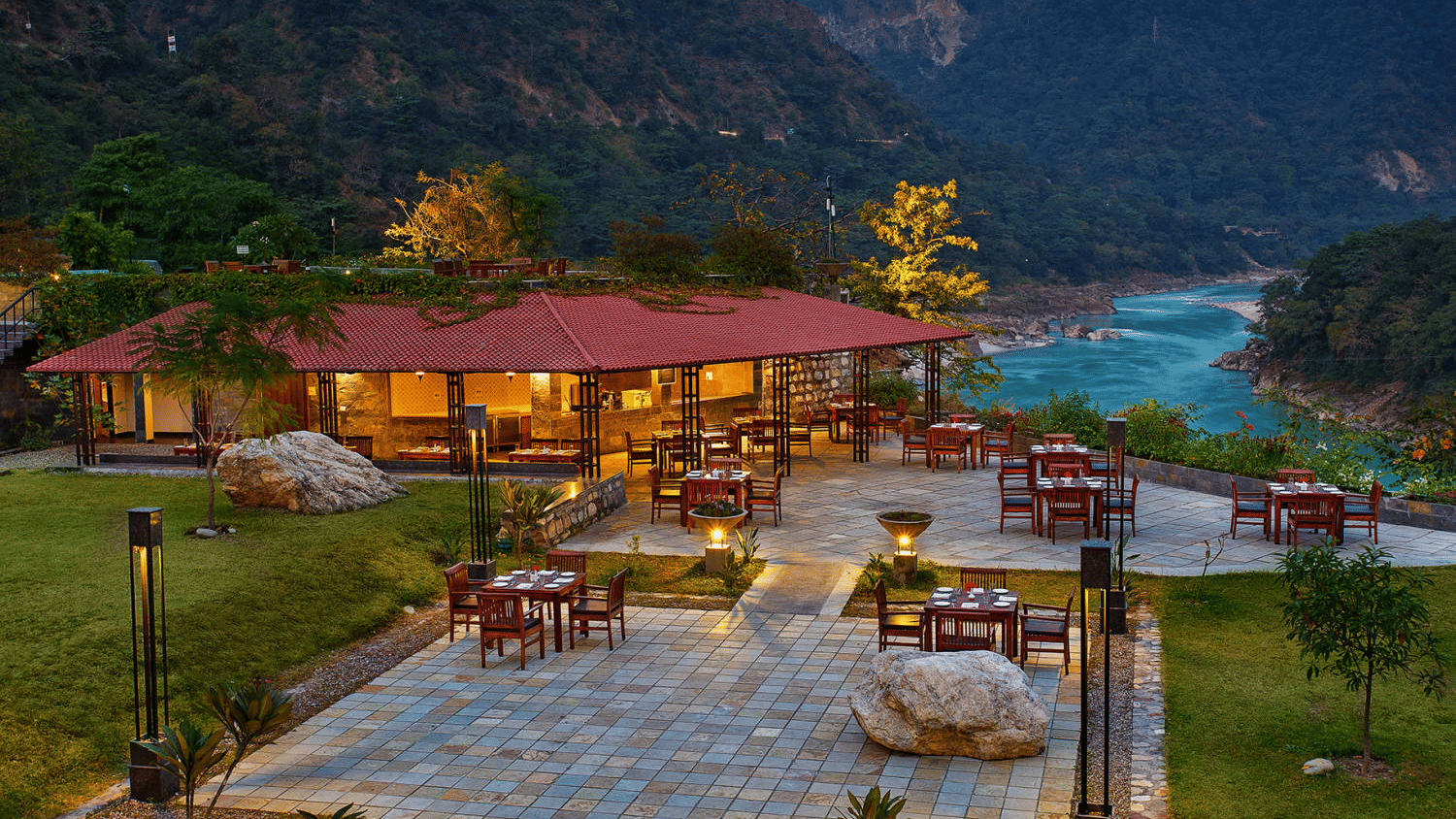 Outdoor dining area by a river, with tables set on a stone patio. A red-roofed pavilion is surrounded by lush greenery and mountains, conveying tranquility - Aloha On the Ganges