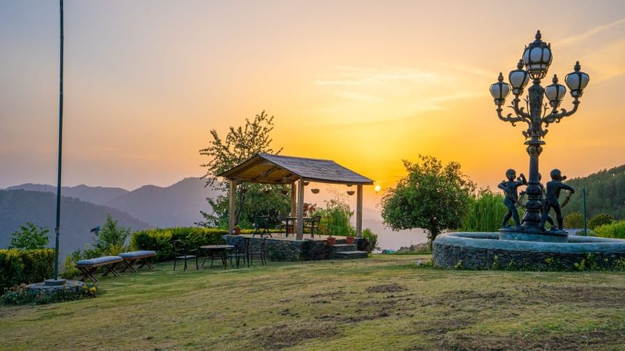 Outdoor view of a landscaped garden with a gazebo and decorative lamp post at sunrise, overlooking hills and distant trees at  The Manor Luxury Apartments, Shimla.