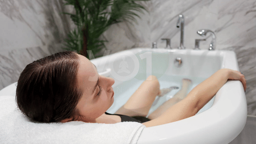 A woman with wet, dark hair relaxing in a white clawfoot bathtub, with water covering her body up to her shoulders at YO1 Longevity & Health Resorts