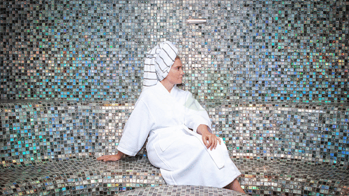 A woman in a white robe and turban-style towel sits on a mosaic tiled bench, looking to her right, in a brightly lit steam room at YO1 Longevity & Health Resorts