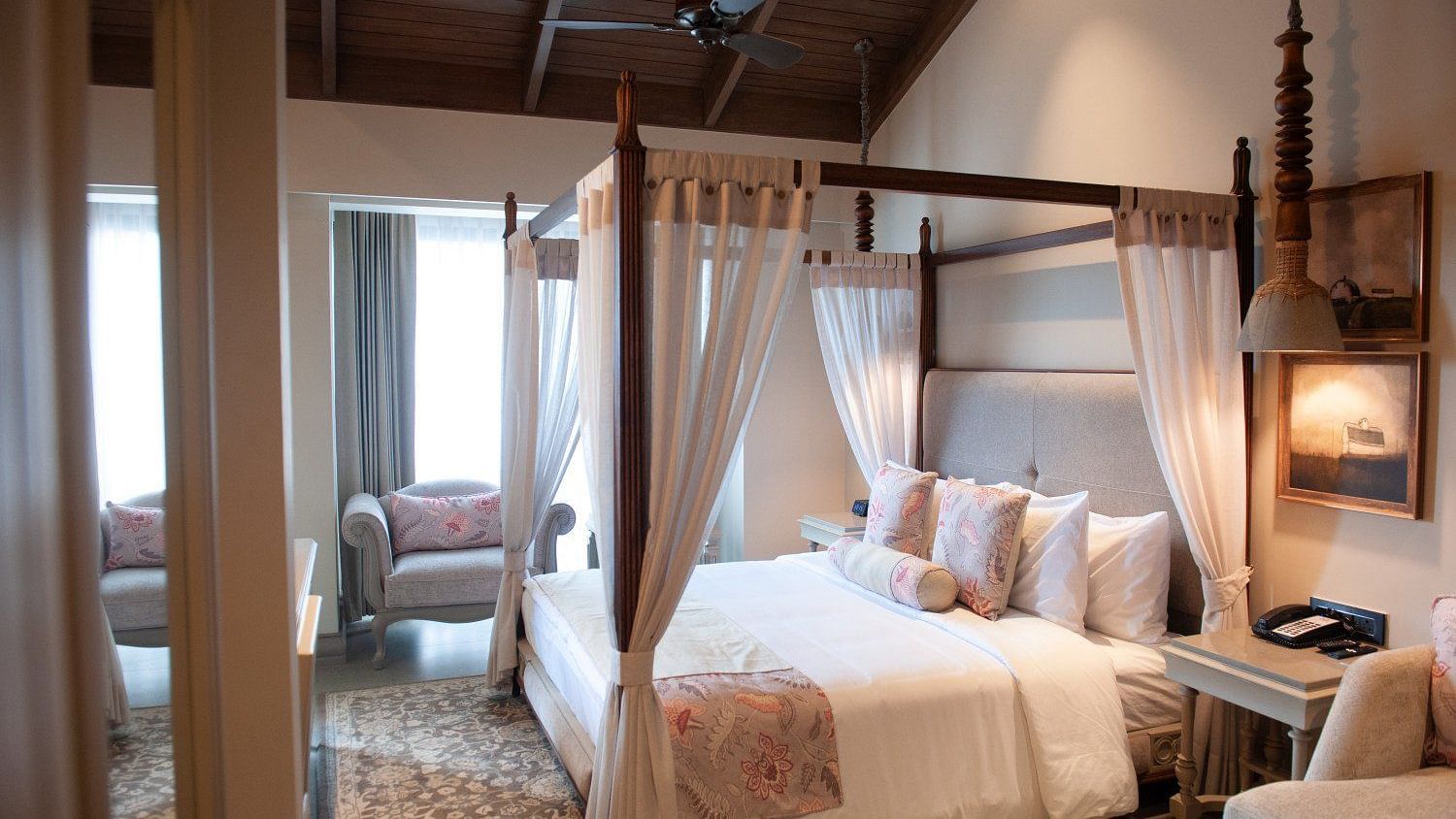  A detailed view of the four-poster bed in an executive room at The Evren, Vagator. The room has a vaulted wooden ceiling, a ceiling fan, and a seating area near the window. The bed is adorned with decorative pillows and curtains.