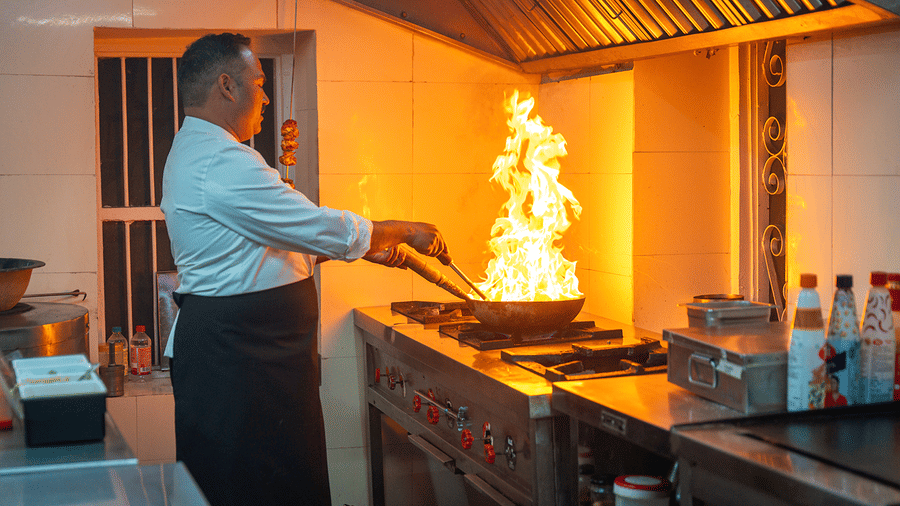 A professional chef managing a high-flame stir-fry in a modern commercial kitchen at Maati Resort By The Lake Hill, Jeolikote.