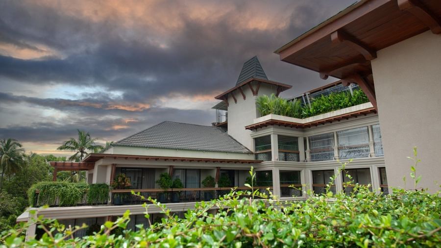 An architectural shot of the exterior of The Evren, Vagator, showing the elegant facade, balconies, and the lush green surroundings. The image is taken from a vantage point that captures the building's design against a dramatic sky.