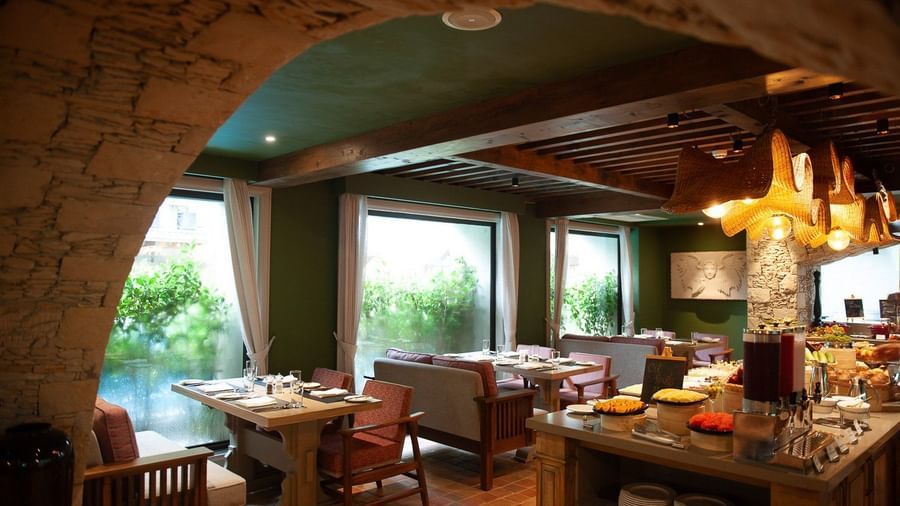 A wide-angle view of the dining area at the Mila Restaurant at The Evren, Vagator, from the corner of the stone archway featuring wooden tables, comfortable chairs and banquettes, and a buffet station with a variety of food.