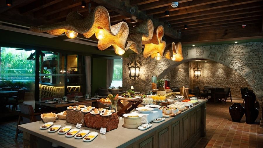 A lavish breakfast buffet spread at Mila Restaurant at The Evren, Vagator, featuring a variety of dishes on a large central island. The restaurant has a rustic yet modern ambiance with woven light fixtures, a stone archway, and a view of the outdoors.