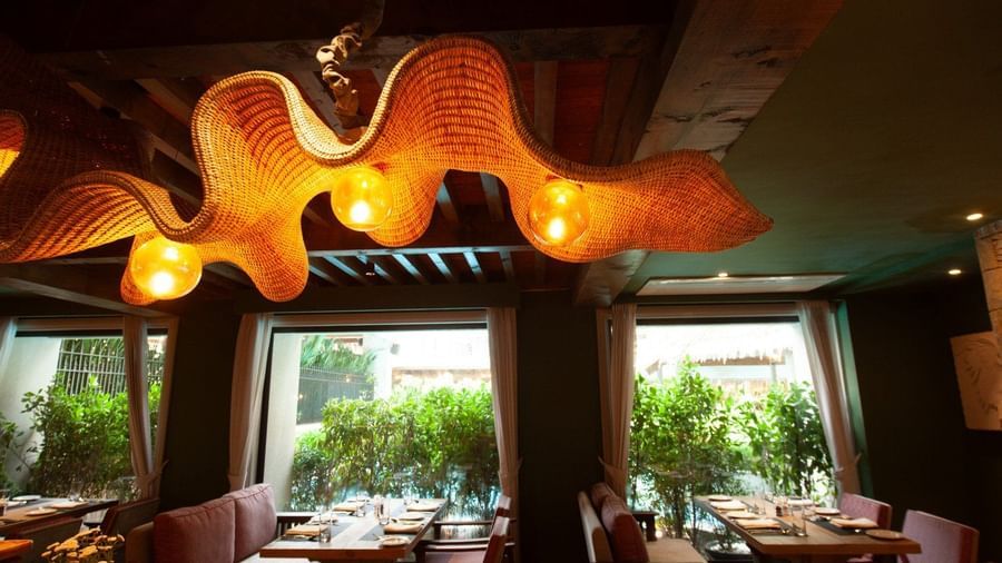 A detailed shot of the interior of Mila Restaurant at The Evren, Vagator, highlighting the unique, woven light fixtures hanging from the wooden ceiling. The restaurant has large windows with a view of greenery and comfortable seating.