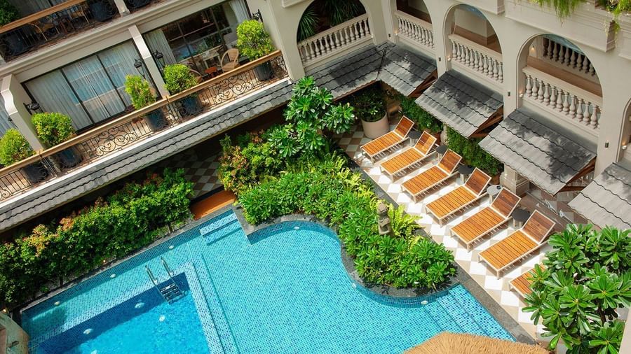 Top-view shot of a bright swimming pool tiled with blue mosaic tiles surrounded by lush green bushes.