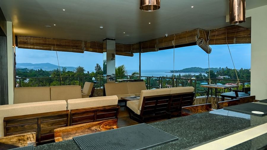 An outdoor balcony seating area with light-coloured chairs and a table, offering a panoramic view of lush green hills and a distant body of water - Sea Hills Hotels, Sri Vijaya Puram (Port Blair)