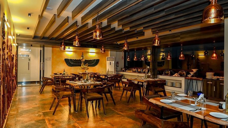 A wide view of a dimly lit restaurant with wooden tables and chairs, warm lighting, and unique wall murals depicting figures - Sea Hills Hotels, Sri Vijaya Puram (Port Blair)