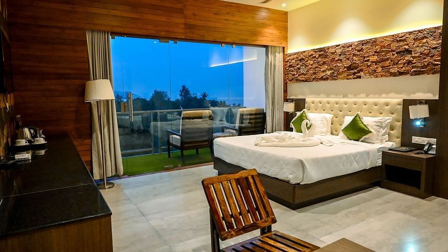 A hotel bedroom with a large bed, a comfortable armchair, and a wide view of the outdoors through a large glass door leading to a balcony - Sea Hills Hotels, Sri Vijaya Puram (Port Blair)