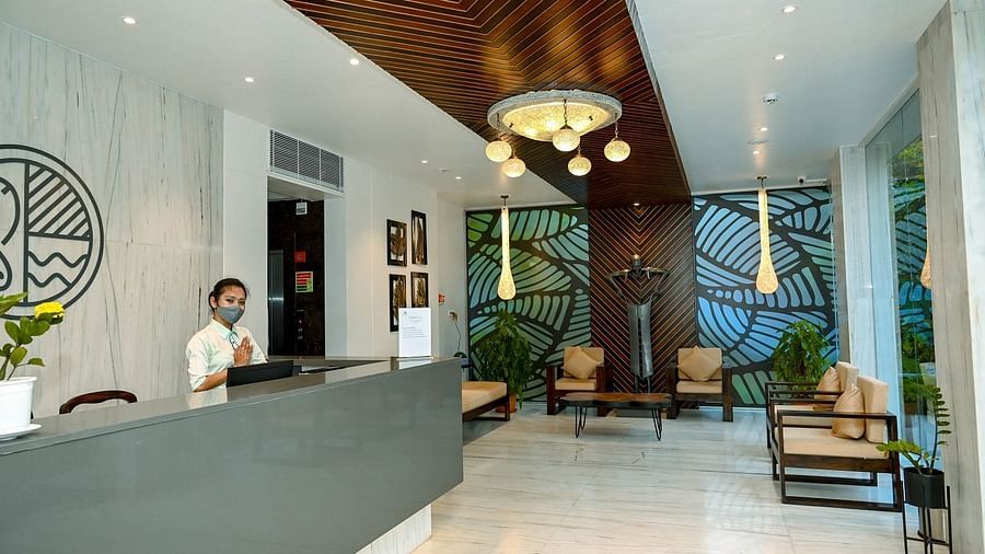 A view of a hotel reception desk and lobby area, showing the minimalist design, seating, and an attendant at the counter - Sea Hills Hotels, Sri Vijaya Puram (Port Blair)