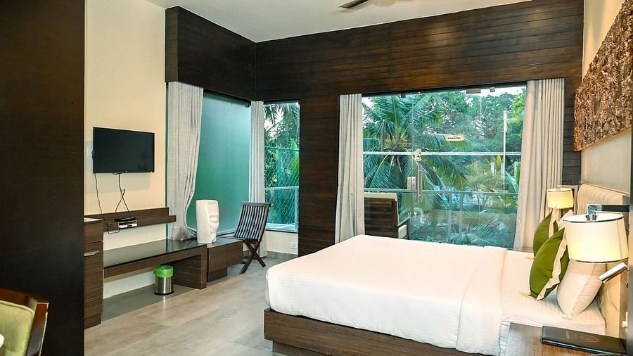 A hotel room with a large bed, a television, and a panoramic window revealing a vibrant green landscape outside, under a ceiling fan - Sea Hills Hotels, Sri Vijaya Puram (Port Blair)
