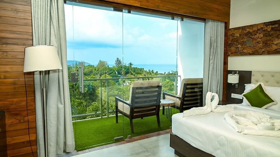 A bright hotel room with a large bed, a comfortable chair on a private balcony, and a panoramic view of the tropical landscape through a large glass door - Sea Hills Hotels, Sri Vijaya Puram (Port Blair)