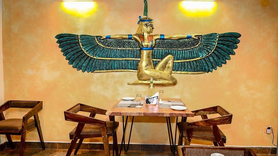 A small, intimate dining setting in a restaurant with two chairs and a table, positioned in front of a striking wall decoration of a winged figure in blue and gold - Sea Hills Hotels, Sri Vijaya Puram (Port Blair)