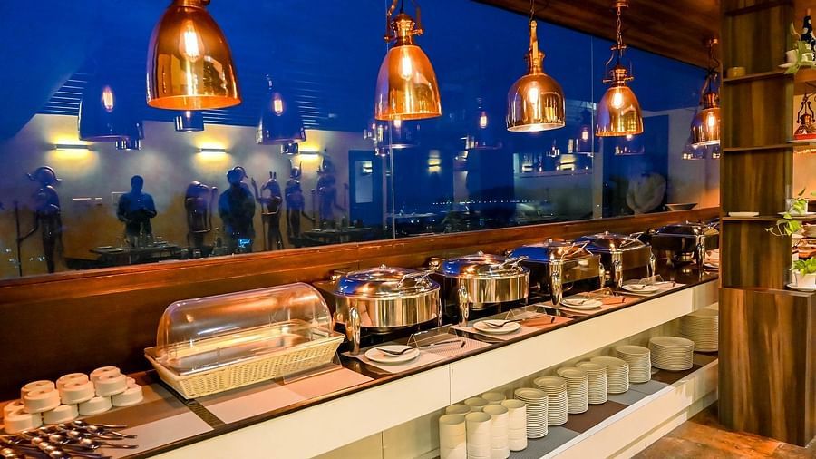 A buffet area in a restaurant, with various dishes in chafing dishes, glasses, and utensils, illuminated by warm hanging lights - Sea Hills Hotels, Sri Vijaya Puram (Port Blair)