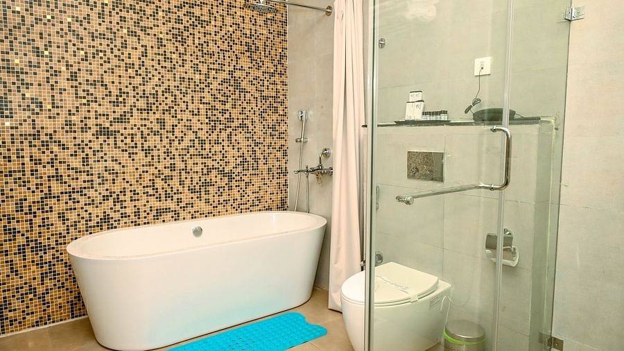 A modern bathroom with a freestanding white bathtub, a toilet, and light-coloured mosaic tiles on the walls, with a blue bathmat on the floor - Sea Hills Hotels, Sri Vijaya Puram (Port Blair)