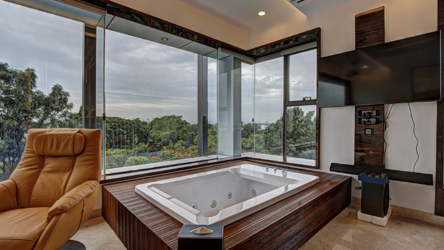 A Jacuzzi tub at the Grand Suite alongside a reclining arm chair at SRS Suites, Koramangala, overlooking the lush green views from the window.