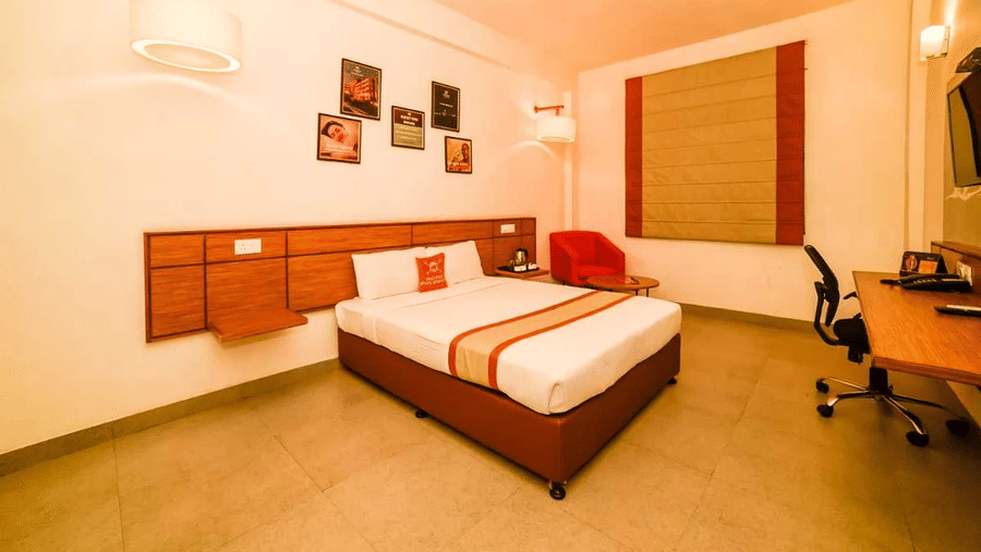 An image of bedroom featuring a queen size bed, a work desk and chair, a sofa, and some photo frames on the walls - An image of a reception lobby featuring a reception desk , a flat TV screen, and a seating area for the guest - Max Hotel Prayagraj
