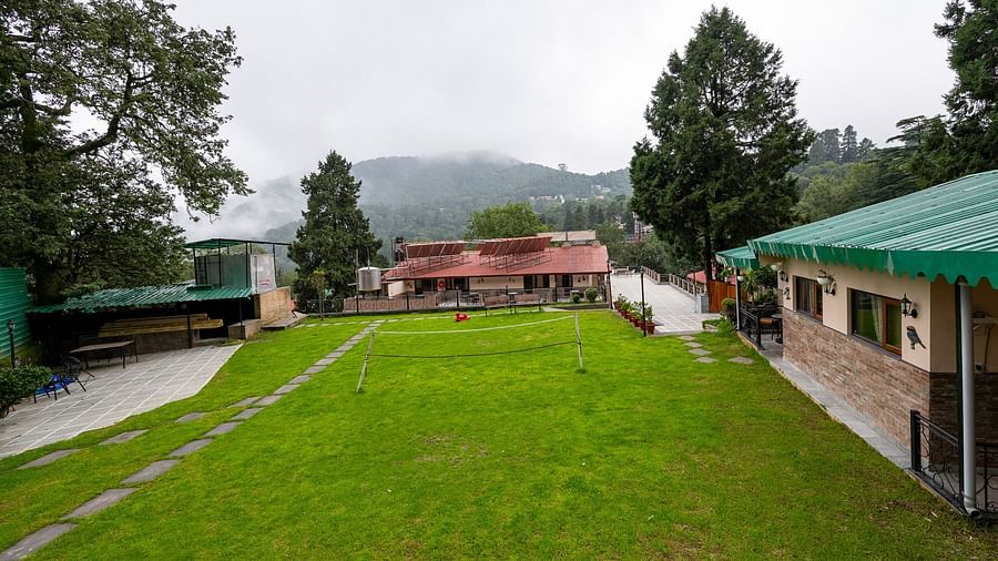 Pristine lawn and grounds at The Pavilion, Nainital with misty mountains in background, green roofed buildings, and well-maintained landscaping.