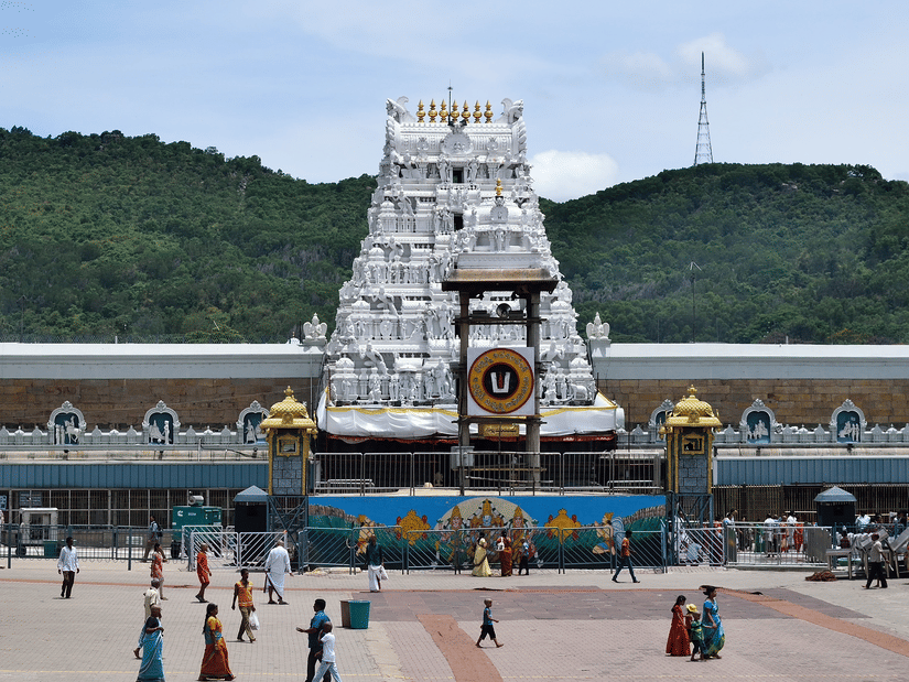 A wide shot of a white, multi-tiered Tirumala Venkateswara Temple tower with golden accents, situated in a courtyard with green hills and a clear sky in the background.