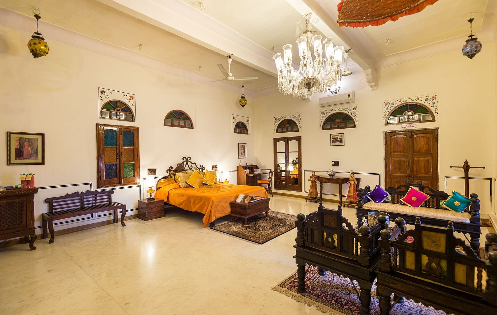 A well-lit bedroom at Fort Barli, featuring a vibrant orange bedspread, a crystal chandelier, and antique furniture, offers a spacious and elegant retreat.