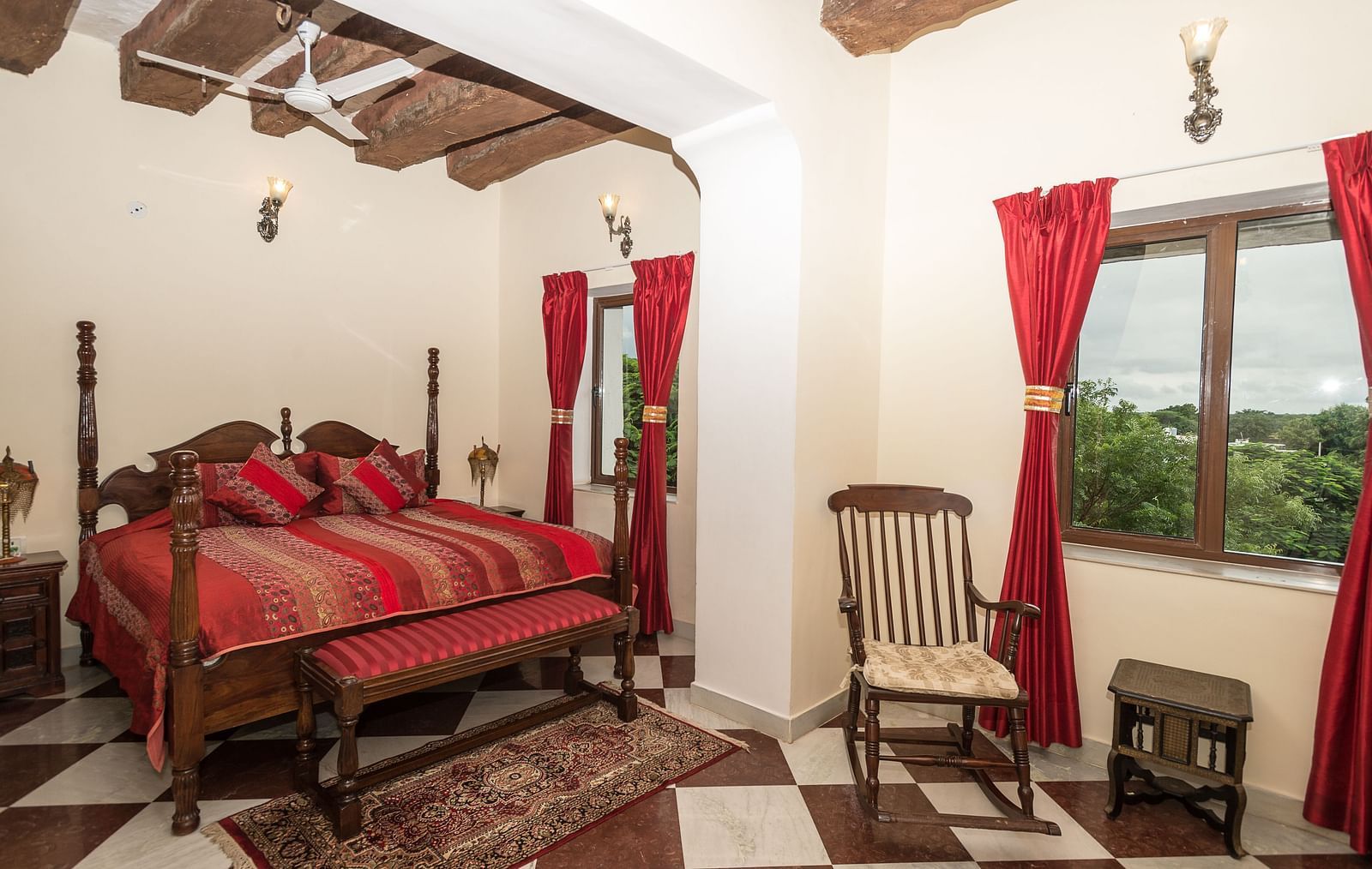 A spacious bedroom at Fort Barli, featuring a four-poster bed with red accents, checkered floors, and exposed wooden beams, offers a blend of rustic charm and elegance.