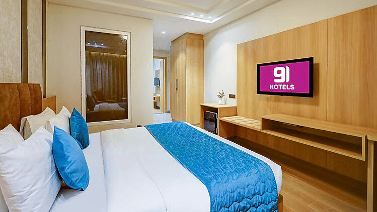 A bedroom at Hotel 91 HUDA Premier, Gurugram with a king size bed, pillows, headboard, wall-mounted TV, desk, and seating chair.