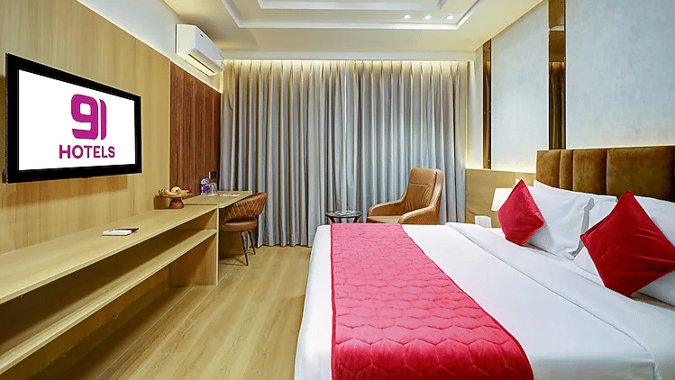A bedroom at Hotel 91 HUDA Premier, Gurugram with a king size bed, runner, pillows, bedside table, wall-mounted TV, desk, and armchairs.