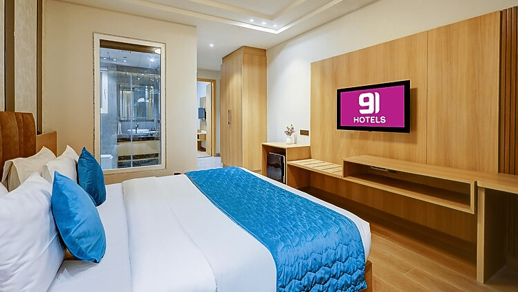 A bedroom at Hotel 91 HUDA Premier, Gurugram with king size bed, wall-mounted TV unit, wooden panel, and door leading to balcony or window area.