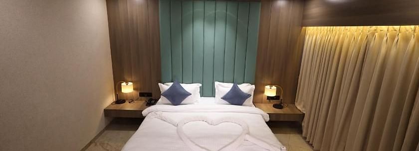 The image shows a hotel bedroom with a bed featuring a heart-shaped design, bedside tables with lamps, and curtains - Lords Eco Inn Navsari.