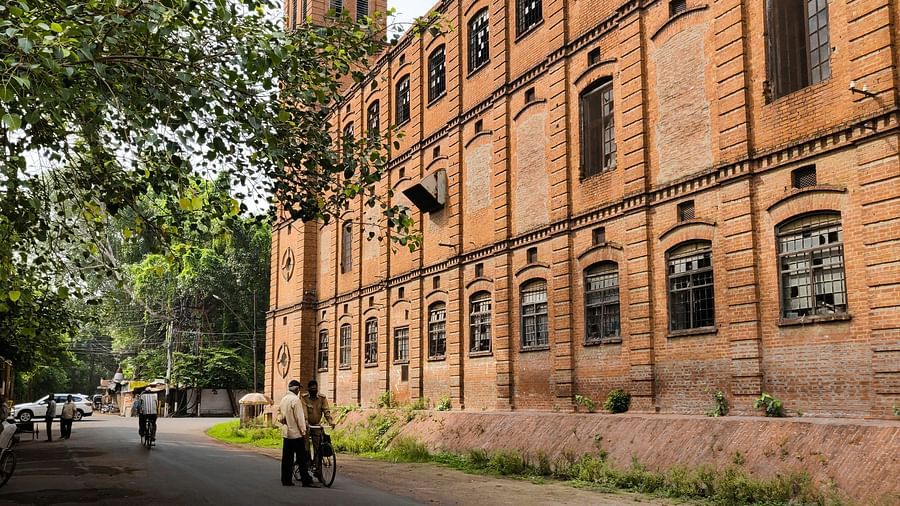 An image of a brick building next to some trees and there is a road between on which people are walking