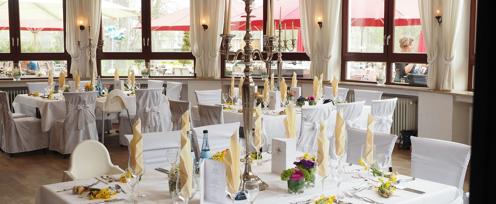 A banquet hall with tables covered with white linens and adorned with floral arrangements, candles, and elegant place settings.