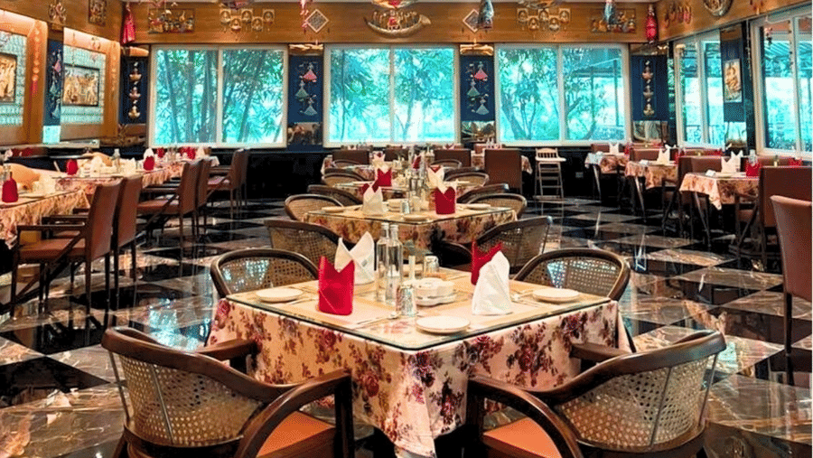 Rangmahal restaurant at MAYFAIR Tea Resort, Siliguri features tables and chairs arranged across a floor. The ceiling contains recessed panels with lighting fixtures and decorations that hang above the tables. 