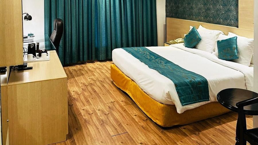 A cosy hotel room at Amer Greens featuring a large bed, hardwood floors, an accent wall, and a large window covered by floor-to-ceiling drapes. Room