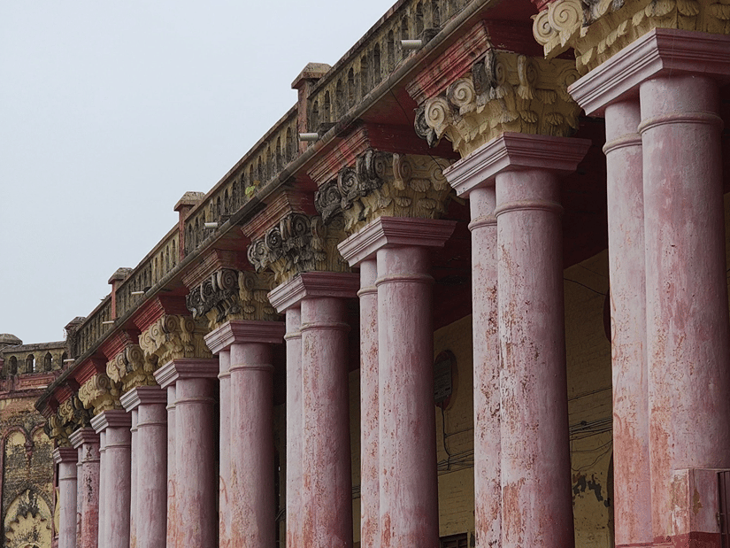 Stone colonnade with carved pillars along an outdoor pink balustrade, set against an overcast sky.