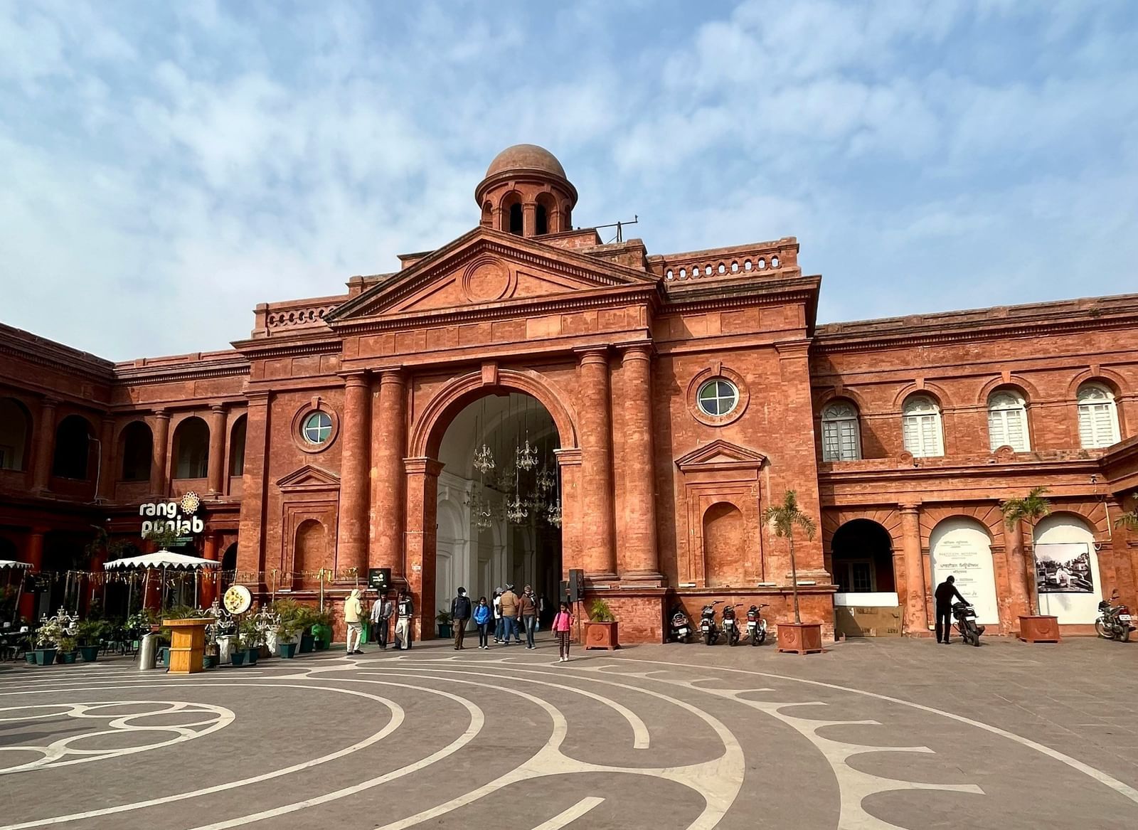 Historic red brick building with an arched entrance, surrounded by a cobbled plaza with a geometric pattern.