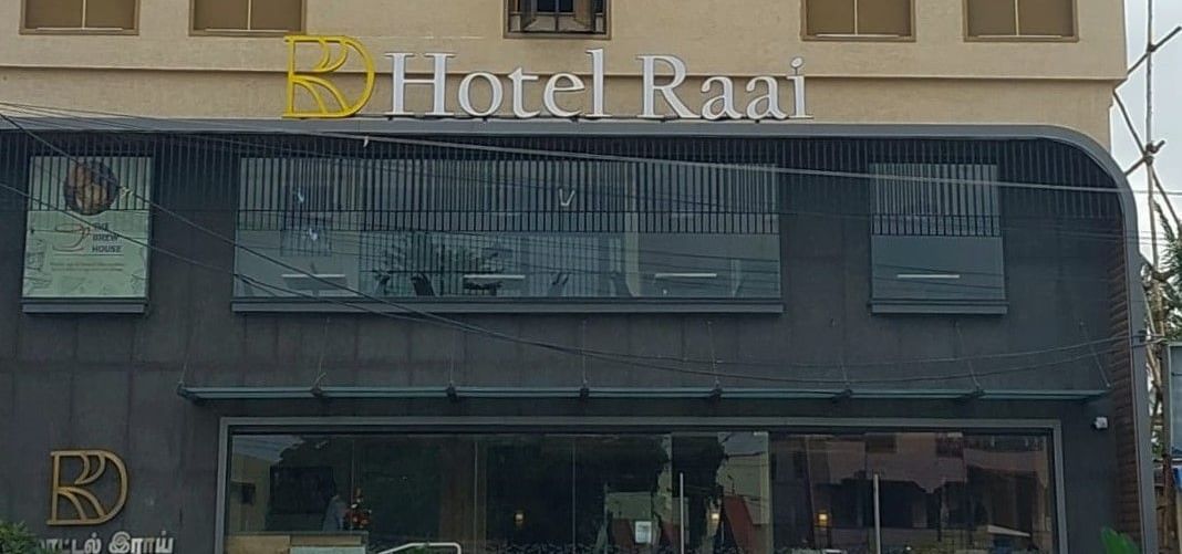 Facade of Hotel Raai, Hosur featuring four storey building. The ground floor has glass walls at the entrance and the other floors have concrete walls with long windows facing the street.