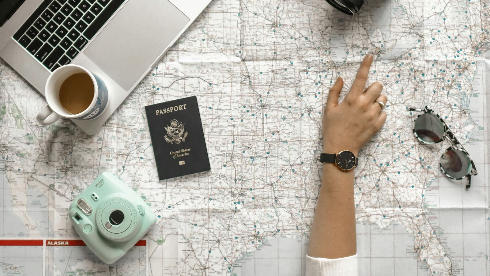 A person pointing at a map with a laptop, camera, passport, instant camera, and sunglasses laid out.