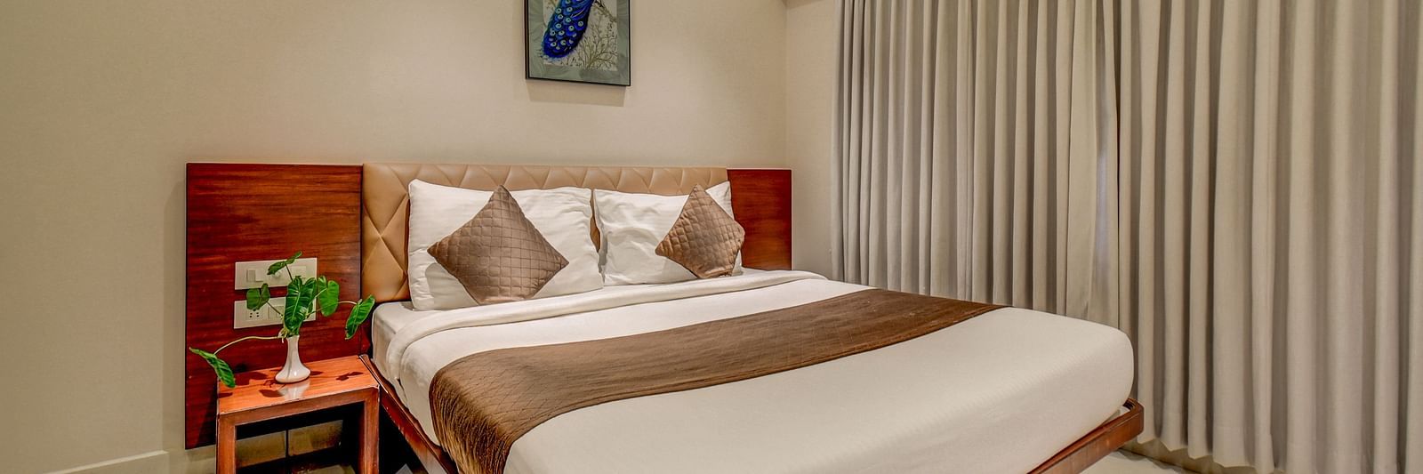 A spacious room at Harigovinda's JNStay with a large bed, a bedside table, large windows fully covered with curtains, and a wall-mounted photo frame above the headboard